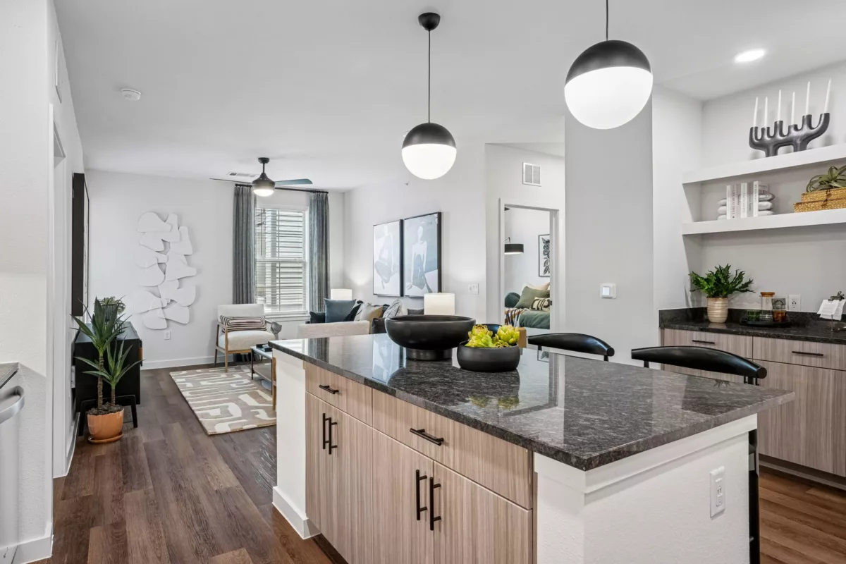 Open-concept kitchen with granite island, pendant lights, and wood-look cabinets, overlooking a living room with white walls and gray curtains.
