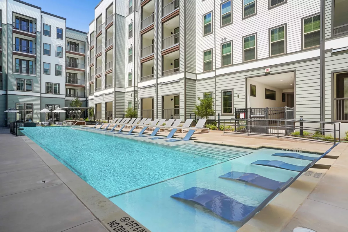 Outdoor pool with lounge chairs in shallow water and on deck, modern building in background.